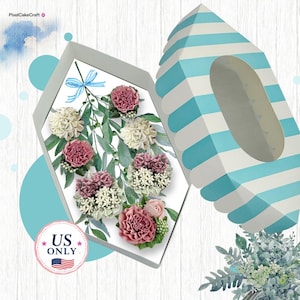 May include: A hexagonal gift box with a light blue and white striped lid, containing a collection of artificial flowers in shades of pink, cream, and white with green foliage. A small blue ribbon bow is attached to the floral arrangement.
