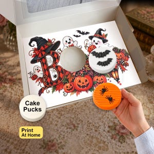 May include: A white cardboard box containing a Halloween-themed insert with a large letter B, ghosts, pumpkins, and bats. One white circular cake puck and one orange circular cake puck with two black spider decorations are visible.