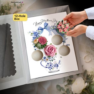 May include: A white 12-hole box insert featuring a printed design of a blue teapot, blue ribbons, and various floral arrangements. The design includes the text Happy Mother's Day in black script. A hand places a pink floral cupcake into the box.