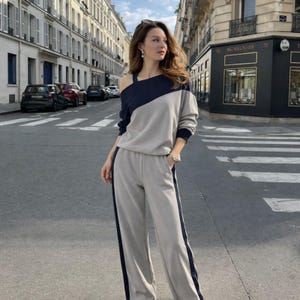 May include: A two-piece athleisure set in gray and navy blue. The top is an asymmetrical long-sleeve sweatshirt with a diagonal color block design. The pants are wide-legged with a navy blue stripe down the side. The outfit is worn outdoors.