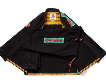 Puede incluir: Un gi de artes marciales negro con un parche color verde azulado con el texto rojo GARFIELD en la parte delantera. El forro interior del cuello tiene un patrón geométrico amarillo y negro. Parches adicionales con texto rojo están en las mangas.