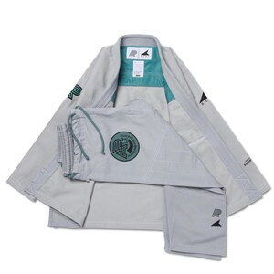Op de afbeelding: Een lichtgrijze vechtsport gi-set bestaande uit een jas en een bijpassende broek. De jas heeft teal-kleurige binnenpanelen en zwarte geborduurde logo's. De broek is voorzien van een trekkoord en een ronde patch met de letters AP in zwart.