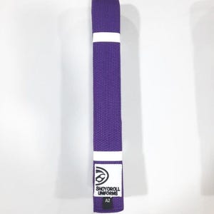 Może przedstawiać: Fioletowy pas do sztuk walki z białym paskiem i etykietą z napisem "SHOYOROLL UNIFORMS" i "A2". Pas jest wykonany z tkanej tkaniny i jest przeznaczony do treningu i zawodów w sztukach walki.