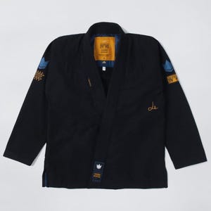 Puede incluir: Uniforme negro de artes marciales con detalles azules y dorados. El uniforme presenta detalles bordados en los hombros y el pecho, junto con una etiqueta amarilla cerca del cuello. Las mangas son largas y el uniforme está plano.