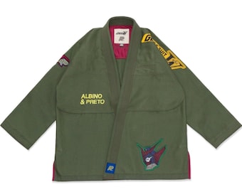 Puede incluir: Gi de artes marciales verde oliva con el texto "ALBINO & PRETO" en la parte delantera. El gi presenta parches con diseños gráficos y el texto "G-SUIT". El forro interior es de color magenta oscuro.