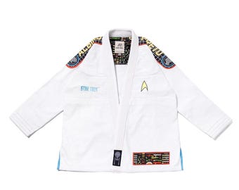Puede incluir: Un kimono de artes marciales blanco con temática de Star Trek, con una insignia amarilla de la Flota Estelar en el pecho, texto Star Trek en azul y parches coloridos del espacio en los hombros y el dobladillo. El forro tiene un patrón de rejilla.