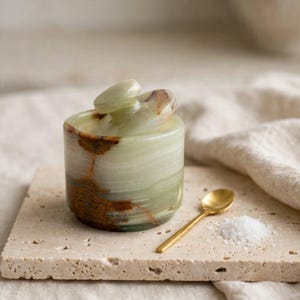 Puede incluir: Un recipiente cilíndrico de piedra verde claro con tapa a juego, que presenta vetas marrones prominentes en toda la superficie pulida. Una pequeña cuchara de metal dorado descansa junto al recipiente, acompañada de un poco de sal blanca.