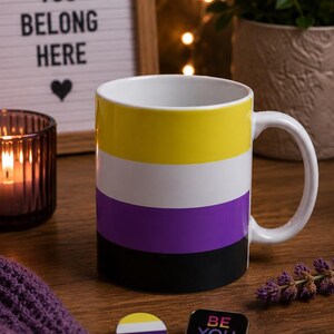 May include: A white ceramic mug featuring horizontal stripes in yellow, white, purple, and black. Beside the mug are two small pins: one circular pin with the same yellow, white, purple, and black stripes, and one square pin with the text BE YOU.