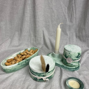 Könnte beinhalten: Eine Sammlung dekorativer Artikel in Grün- und Weißtönen. Dazu gehören ein Kerzenhalter, eine kleine runde Kerze, ein Behälter mit Erdnüssen und zwei Behälter mit Deckel. Die Artikel sind auf einem grauen Stoff angeordnet.