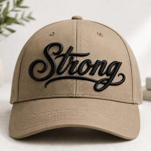 May include: A tan baseball cap featuring the word Strong in raised black script lettering across the front. The cap has a structured crown, a curved brim, and stitched eyelets on the top panels, providing a clean and simple design.