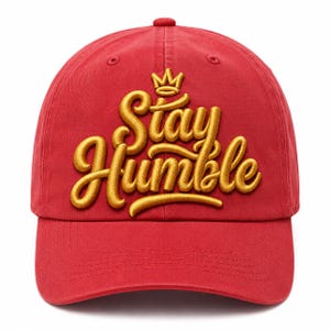 May include: A red baseball cap featuring the embroidered gold text Stay Humble in a cursive script font. Above the text is a small gold embroidered crown icon. The cap has a structured front panel, a curved brim, and matching red stitching.
