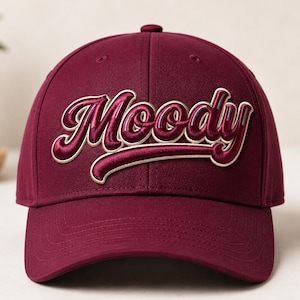 Puede incluir: Una gorra de béisbol de color burdeos oscuro con la palabra Moody bordada en relieve en una fuente de estilo manuscrito en la parte delantera. Las letras están delineadas en un color crema claro, creando un contraste con la tela oscura.