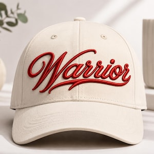 May include: A cream-colored baseball cap featuring the word Warrior embroidered in a bold, raised red script font across the front panels. The cap includes a curved brim, a button on the top crown, and stitched eyelets for ventilation.