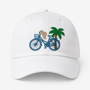 May include: A white baseball cap featuring an embroidered design of a blue bicycle with a front basket, a tan straw hat hanging from the handlebars, and a green palm tree with a brown trunk positioned behind the bicycle frame.