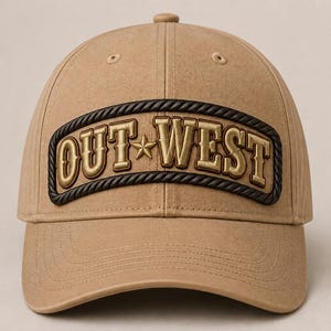 May include: A tan baseball cap featuring a central patch with the text OUT WEST in gold lettering, separated by a small gold star. The patch is framed by a dark brown rope-style border, and the cap has a structured crown with stitched eyelets.