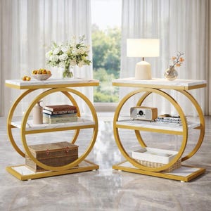 May include: Two matching side tables with a gold metal frame and white marble tops. Each table has a circular design with two shelves. One table has a lamp, while the other has a vase of flowers. The tables are decorated with books, a decorative box, and a small speaker.