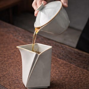 May include: A light gray ceramic tea set with gold trim. Tea is being poured from a lidded teapot into a square-shaped cup. The tea set is on a wooden surface.