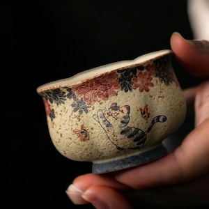 May include: A small, beige ceramic teacup with a whimsical cat illustration. The cup features a floral pattern in red and blue around the rim. The cat is depicted in a playful pose, with a striped body and a curved tail.