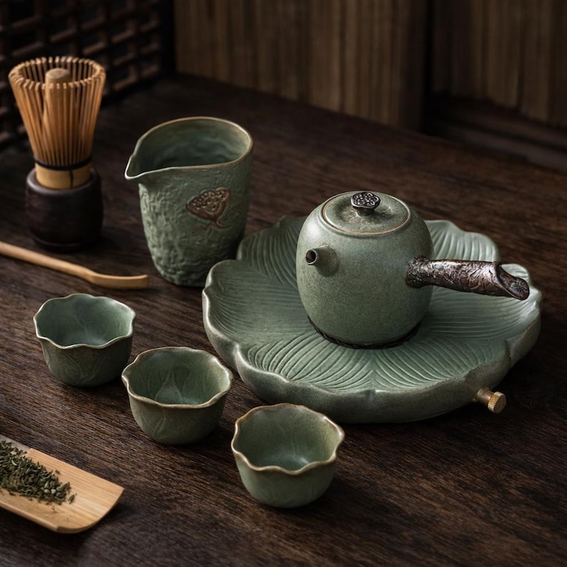 May include: A green ceramic tea set with a teapot, cups, and a serving pitcher. The teapot sits on a leaf-shaped tray. A bamboo whisk and spoon are also present, along with a small wooden tray of tea leaves. The set has a rustic, natural aesthetic.