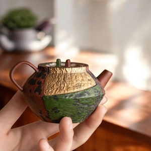 以下が含まれることがあります： ユニークなパッチワークデザインの小さな手作りのティーポット。 ティーポットは、茶色、緑、金などの色の組み合わせで、テクスチャ加工された表面が特徴です。 湾曲したハンドルと小さな注ぎ口があり、お茶を出すのに最適です。
