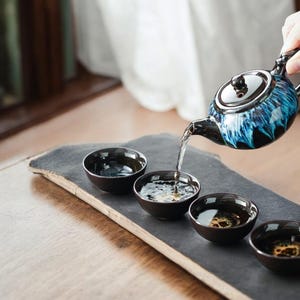 Pode incluir: Uma chaleira de cerâmica com esmalte azul e preto serve chá em pequenas xícaras de chá pretas combinando. O conjunto de chá é disposto em uma bandeja retangular cinza escuro, criando uma apresentação serena e elegante. A chaleira tem uma tampa e uma alça prateadas.