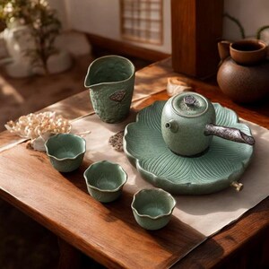 May include: A green ceramic tea set with a teapot, cups, and a serving pitcher. The teapot sits on a leaf-shaped tray. The cups have a scalloped edge. The set is arranged on a wooden table, creating a serene and elegant aesthetic.