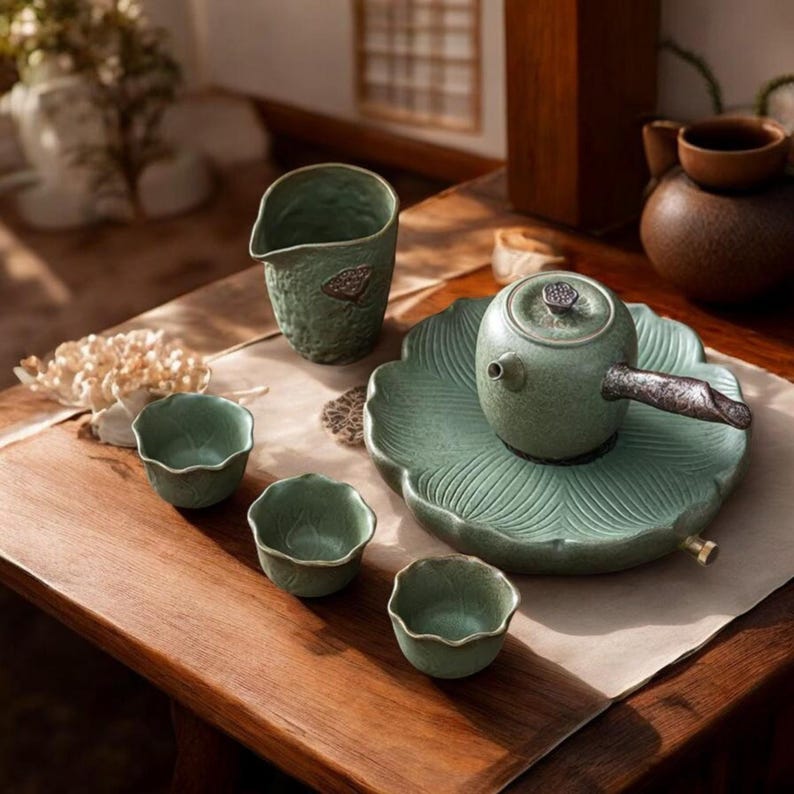 May include: A green ceramic tea set with a teapot, cups, and a serving pitcher. The teapot sits on a leaf-shaped tray. The cups have a scalloped edge. The set is arranged on a wooden table, creating a serene and elegant aesthetic.