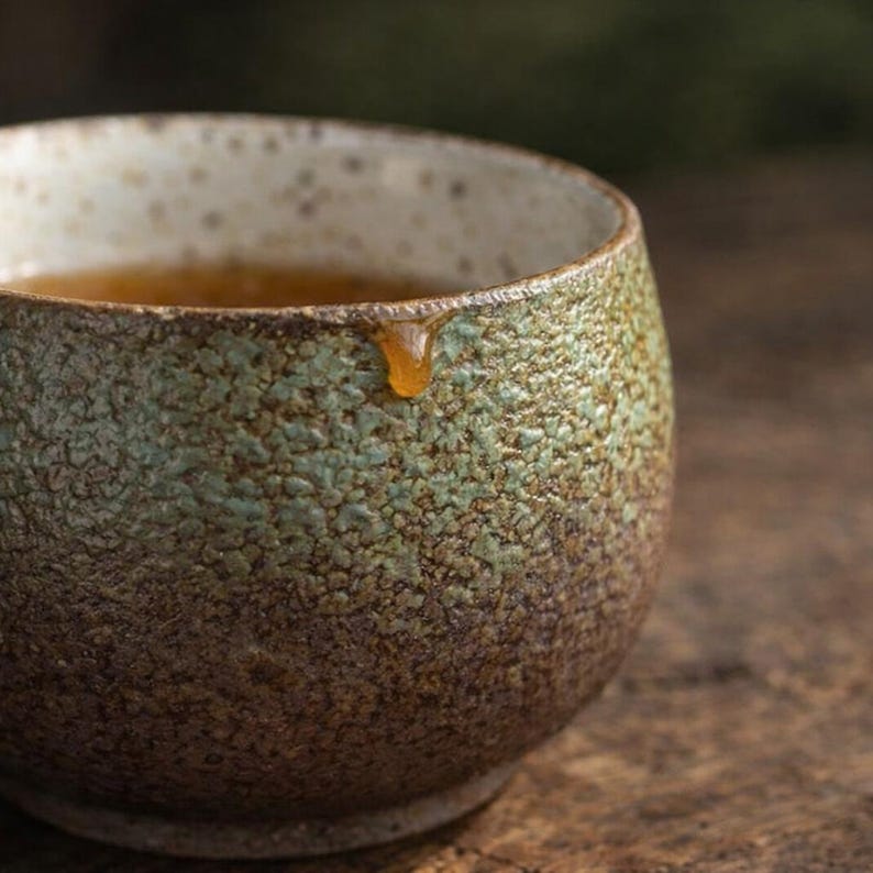 May include: A handcrafted ceramic tea cup with a textured, mottled surface in shades of brown and green. The interior is filled with tea, and a drop of tea hangs from the rim. The cup has a speckled, off-white rim.
