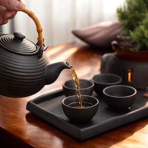 Op de afbeelding: Een zwarte keramische theepot met een bamboe handvat schenkt thee in een kleine zwarte kom. De theeset bevat meerdere kleine kommen op een zwart dienblad. De thee heeft een gouden kleur en er stijgt stoom op uit de kommen.
