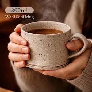 May include: A speckled beige Wabi Sabi mug, filled with a dark beverage, is held by two hands. The mug has a rounded handle and a capacity of 200ml. Steam rises from the warm drink. The mug is a neutral color and has a rustic appearance.