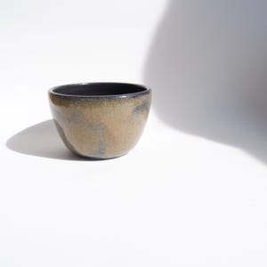 Op de afbeelding: Een kleine, handgemaakte keramische kom met een afgeronde vorm. De buitenkant heeft een gevlekte, aardse kleurenpalet van bruin, bruin en zwart. De binnenkant is effen donkerzwart. De kom staat op een wit oppervlak.