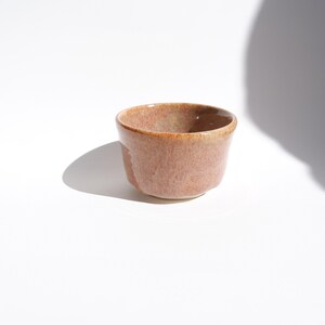 Op de afbeelding: Een kleine, handgemaakte keramische kom met een afgeronde vorm en een getextureerd, gespikkeld oppervlak. De kom heeft een gedempte roodbruine kleur met een lichtere bruine rand. De kom staat op een witte ondergrond.