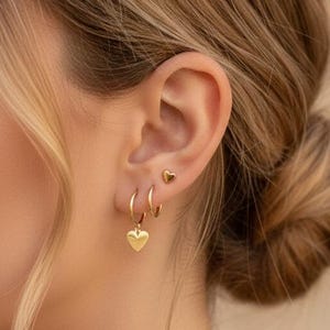 Könnte beinhalten: Nahaufnahme eines Ohrs, das mit drei goldenen Ohrringen geschmückt ist. Die Ohrringe umfassen einen kleinen Herzstecker, einen kleinen Reifen und einen größeren Reifen mit einem Herzanhänger. Die Person hat hellbraunes Haar, das zu einem Dutt frisiert ist.