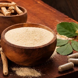 May include: A wooden bowl filled with fine beige powder, accompanied by dried light brown roots, several beige capsules, and green leaves. The arrangement is displayed on a dark wooden surface, showcasing the raw and processed forms of the plant.