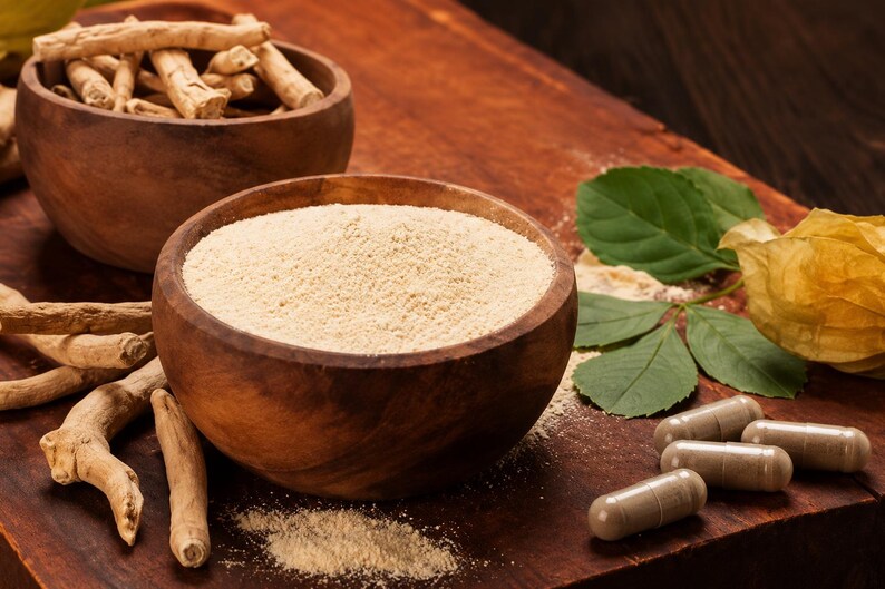 May include: A wooden bowl filled with fine beige powder, accompanied by dried light brown roots, several beige capsules, and green leaves. The arrangement is displayed on a dark wooden surface, showcasing the raw and processed forms of the plant.