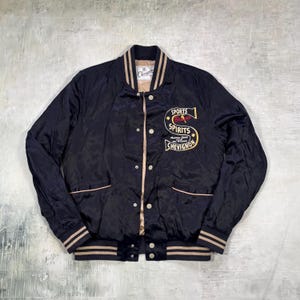 Könnte beinhalten: Schwarze Satin-Bomberjacke mit beigefarbenen und weißen Streifen. Auf der Brust befindet sich ein "SPORTS SPIRITS CHEVIGNON"-Patch. Die Jacke hat einen Druckknopfverschluss und zwei Vordertaschen. Kragen und Manschetten haben beige Streifen.