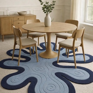 May include: A dining room scene featuring a round wooden table with four chairs. A unique, abstract-shaped rug in shades of blue and navy blue is centered beneath the table. A vase with greenery sits on the table.