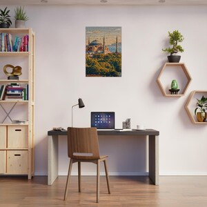 May include: A framed print of a building with multiple domes and minarets, set against a blue sky. The print is displayed on a white wall above a desk with a laptop, lamp, and chair. A wooden bookshelf and decorative shelves with plants are also visible.