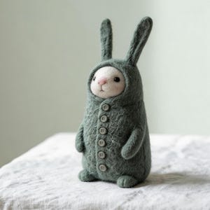 Puede incluir: Figura de conejo de fieltro hecha a mano, vestida con un abrigo verde con capucha y botones. El conejo tiene una cara blanca y una nariz rosa, con orejas largas y erguidas. La figura está sobre una superficie blanca texturizada.