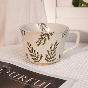 May include: A cream-colored ceramic mug with a handle, adorned with a repeating pattern of green fern-like branches. The mug sits on a white textured surface, with a magazine visible in the foreground.