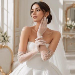 May include: Elegant, sheer white bridal gloves adorned with delicate pearl embellishments. The gloves are long, extending past the elbow, and paired with a strapless white wedding gown. The image captures a classic, romantic bridal look.