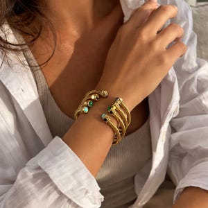 May include: Gold-toned cuff bracelet with a woven design. The bracelet features multiple strands and is adorned with small, colorful gemstones in shades of green, blue, and black. The bracelet is open at the wrist.