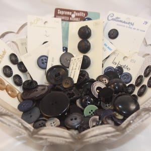 May include: A clear, scalloped glass bowl filled with a variety of buttons in shades of black, brown, and blue. Some buttons are on vintage cards with text like "Supreme Quality" and "Costumakers".