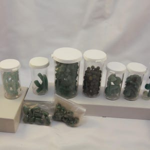 Puede incluir: Surtido de piedras preciosas verdes en frascos de plástico transparente con tapas blancas. Se ven piedras de varias formas y tamaños, incluidas formas redondeadas e irregulares. Algunas piedras están sueltas en bolsas pequeñas. Los frascos están colocados sobre una superficie blanca.