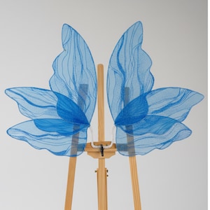 Könnte beinhalten: Ein Paar blaue Schmetterlingsflügel mit einem glitzernden, durchscheinenden Design. Die Flügel werden auf einer hölzernen Staffelei präsentiert, die ihre zarte Struktur und leuchtende Farbe hervorhebt. Ein skurriles Accessoire.
