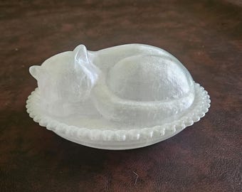 May include: A clear, translucent glass butter dish shaped like a sleeping cat. The lid features the sculpted form of a curled-up feline, while the base is a round, shallow plate with a decorative beaded edge around the entire outer circumference.