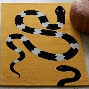 May include: A rectangular mustard yellow area rug featuring a large, winding snake with a black and white patterned body and a smaller, solid black snake with a visible tongue positioned near the bottom right corner of the rug's surface.