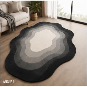 Puede incluir: Una alfombra de forma orgánica irregular que presenta un degradado de bandas onduladas concéntricas en tonos crema, gris claro, gris medio, gris oscuro y negro, creando un efecto de mapa topográfico sobre una superficie de suelo de madera lisa.