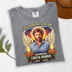 May include: Gray Comfort Colors t-shirt featuring a portrait of Chuck Norris with angel wings, candles, and roses. The text reads "LEGENDS NEVER DIE" and "REMEMBERING CHUCK NORRIS 1940-2024".