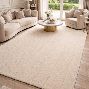 Può includere: Un grande tappeto color crema con un design a coste in un soggiorno. Il tappeto è rettangolare e copre la maggior parte del pavimento. La stanza presenta anche un divano beige, una poltrona e un tavolino rotondo.